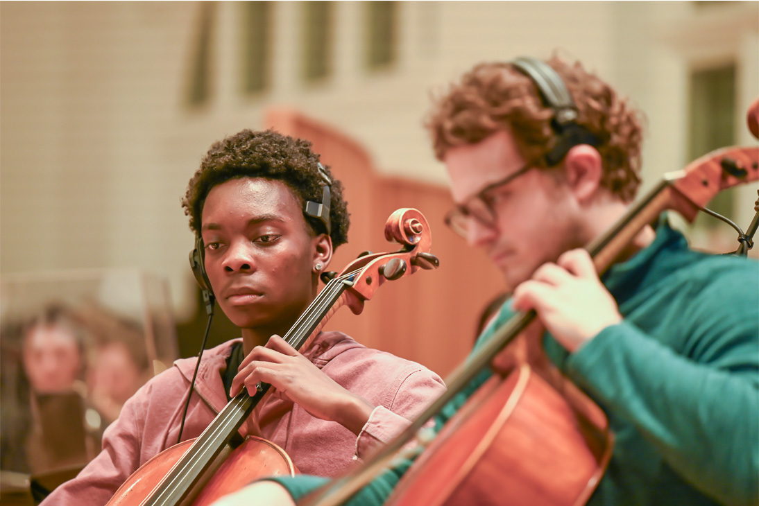 Two male students, holding cellos, rehearsing in the cello section of an orchestra in a Composition for Screen workshop.